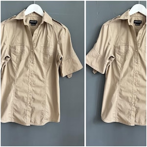 Può includere: Camicia beige a maniche corte con due tasche sul petto e spalline, appesa a una gruccia bianca. La camicia ha un colletto classico e una vestibilità su misura. Adatta per un abbigliamento casual o semi-formale, è un capo versatile.