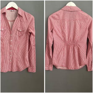 以下が含まれることがあります： 赤と白のストライプのボタンダウンシャツ。長袖、襟付き、胸ポケットが2つ付いています。白いハンガーにかけられており、背景はグレーです。シャツの背面も表示されています。