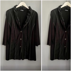 May include: Black long-sleeve jacket with a ruffled collar and white trim. The jacket has three buttons and is hanging on a white hanger. The jacket is a dark color and appears to be made of a lightweight material.