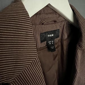 K&ouml;nnte beinhalten: Ein brauner Blazer mit wei&szlig;en Streifen und einem schwarzen H&M-Etikett. Das Etikett zeigt "EUR 40 US 10" und "Hergestellt in China".
