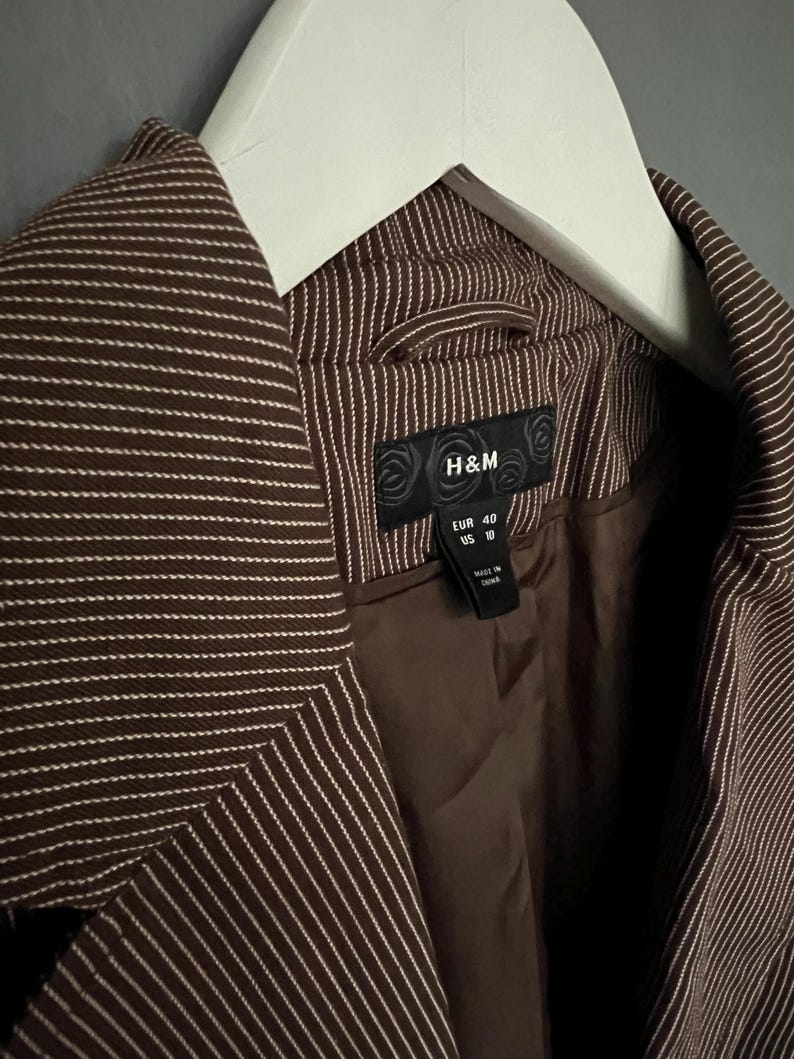 K&ouml;nnte beinhalten: Ein brauner Blazer mit wei&szlig;en Streifen und einem schwarzen H&M-Etikett. Das Etikett zeigt "EUR 40 US 10" und "Hergestellt in China".