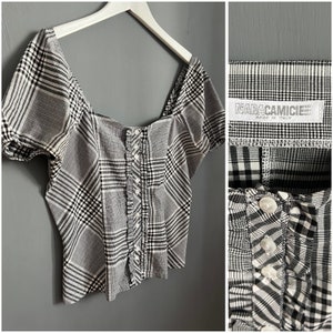 May include: A black and white plaid button-up top with a square neckline and short puff sleeves. The top has a ruffle trim down the front and a label that reads "MARACAMICIE Made in Italy".