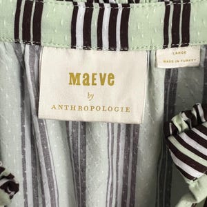 May include: A green and white striped shirt with a brown stripe detail. The shirt has a ruffled neckline and a label that reads "Maeve by Anthropologie" and "Large, Made in Turkey".