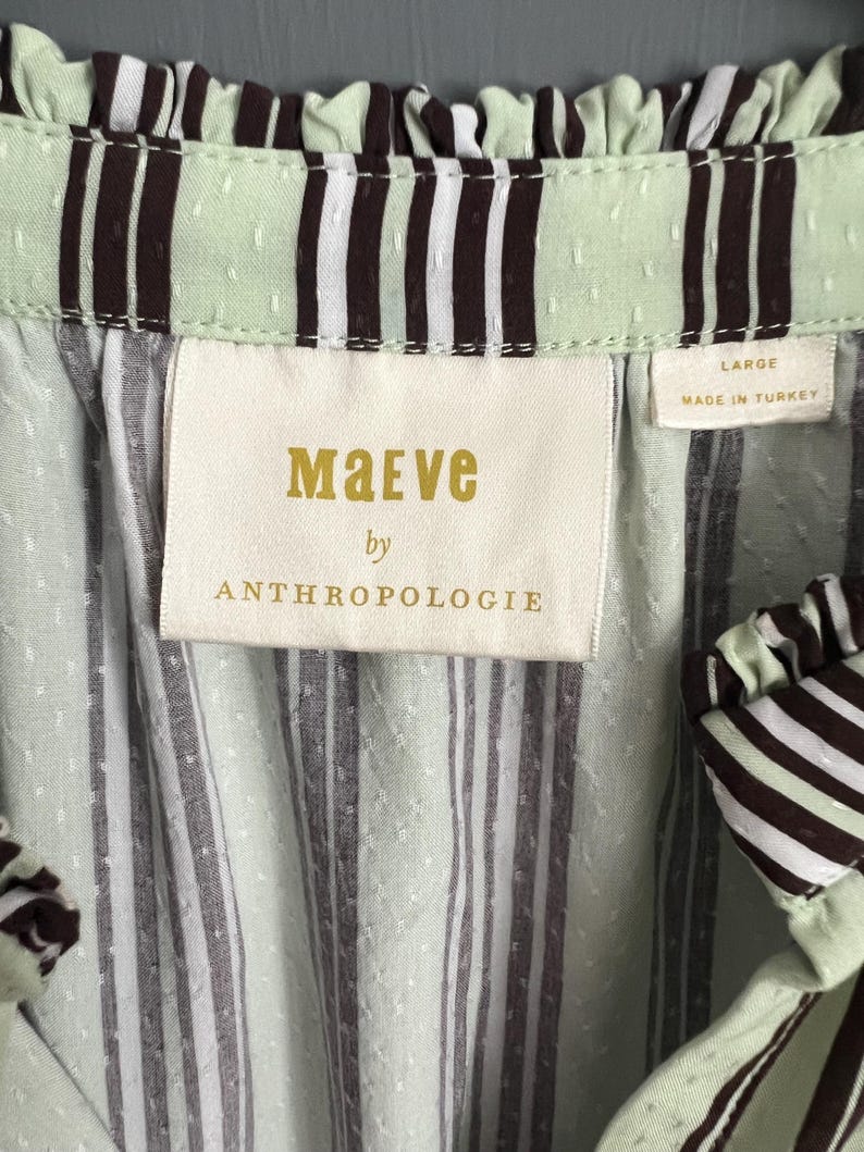 May include: A green and white striped shirt with a brown stripe detail. The shirt has a ruffled neckline and a label that reads "Maeve by Anthropologie" and "Large, Made in Turkey".