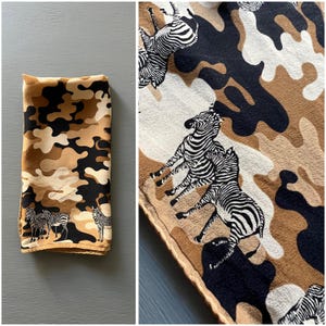 May include: A rectangular scarf featuring a camouflage pattern in shades of tan, black, and cream. The fabric is decorated with a repeating print of black and white zebras standing in various positions across the camouflage background.