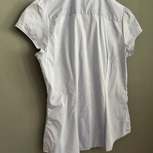 Op de afbeelding: Lichtblauw overhemd met korte mouwen en een subtiel streepjespatroon. Het overhemd heeft een klassieke kraag en een getailleerde pasvorm. De achterkant toont het getailleerde ontwerp en de textuur van de stof. Het overhemd hangt aan een witte hanger.