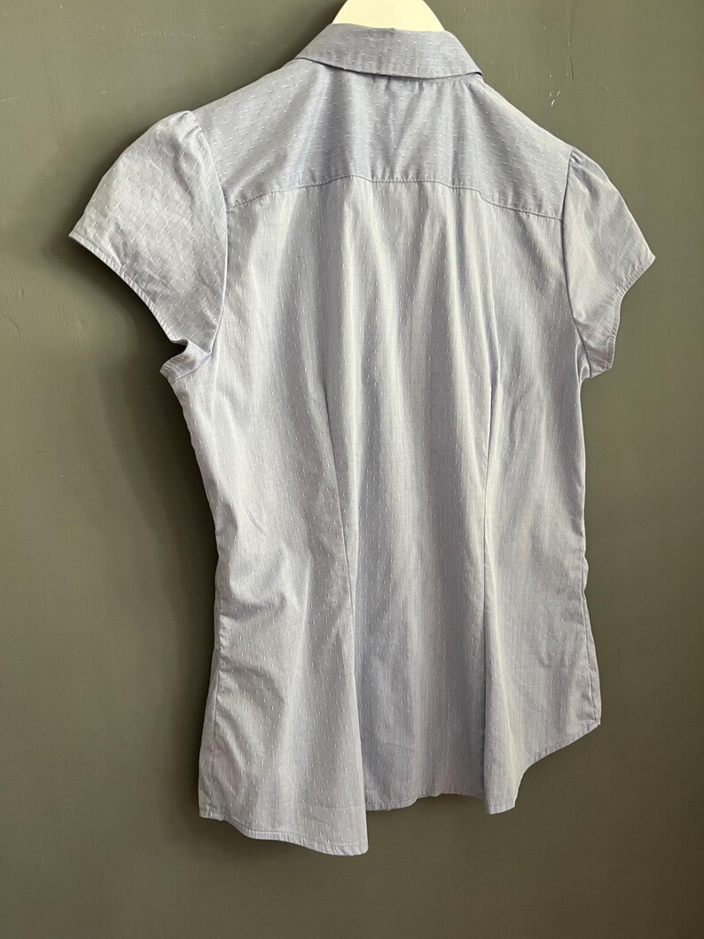 Op de afbeelding: Lichtblauw overhemd met korte mouwen en een subtiel streepjespatroon. Het overhemd heeft een klassieke kraag en een getailleerde pasvorm. De achterkant toont het getailleerde ontwerp en de textuur van de stof. Het overhemd hangt aan een witte hanger.