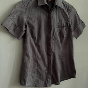 May include: A charcoal grey short-sleeved button-down shirt with vertical stripes. The shirt has a collared neckline, a chest pocket, and cuffed sleeves. The shirt is hanging on a white hanger.