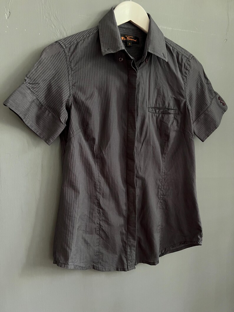 May include: A charcoal grey short-sleeved button-down shirt with vertical stripes. The shirt has a collared neckline, a chest pocket, and cuffed sleeves. The shirt is hanging on a white hanger.