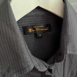 May include: A close-up of a charcoal grey button-down shirt with a vertical striped texture, hanging on a white hanger. The label reads "THE ORIGINAL Ben Sherman" with a size "S".