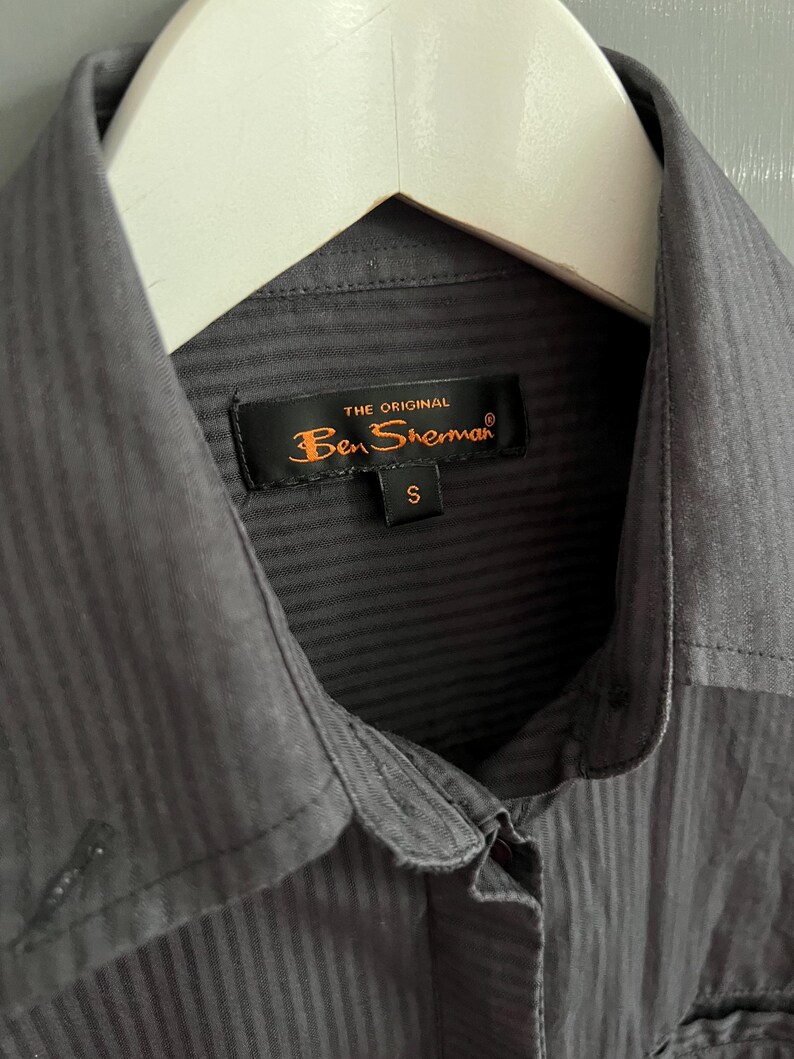 May include: A close-up of a charcoal grey button-down shirt with a vertical striped texture, hanging on a white hanger. The label reads "THE ORIGINAL Ben Sherman" with a size "S".