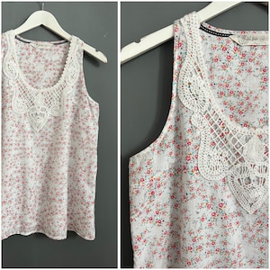 May include: White sleeveless tank top with a floral print and a white crochet trim around the neckline. The top has the text "Lil Luxe" on the shoulder.