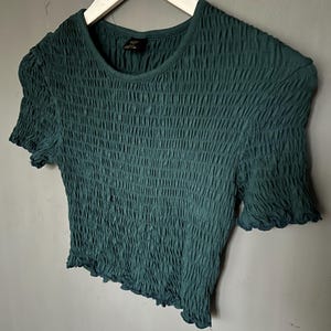 K&ouml;nnte beinhalten: Ein dunkel t&uuml;rkisfarbenes Oberteil mit kurzen &Auml;rmeln und einem strukturierten, ger&uuml;schten Design. Das Shirt hat einen runden Ausschnitt und ger&uuml;schte Kanten an den &Auml;rmeln und am Saum. Es h&auml;ngt an einem wei&szlig;en Kleiderb&uuml;gel vor grauem Hintergrund.