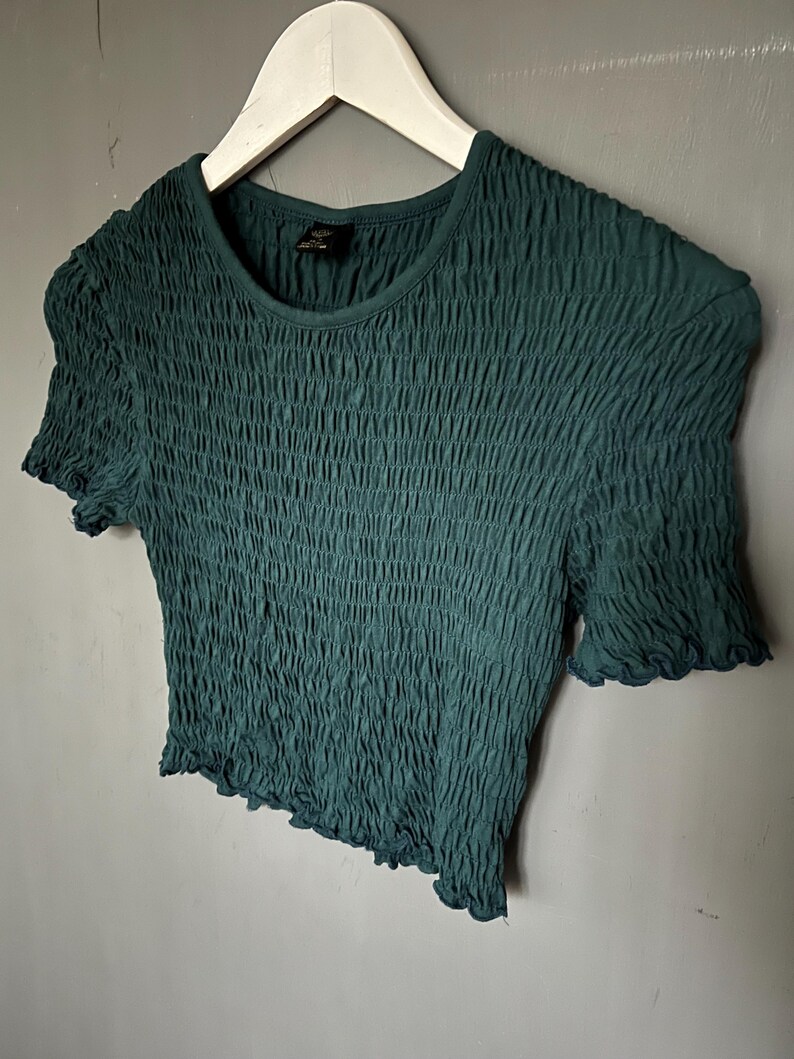 K&ouml;nnte beinhalten: Ein dunkel t&uuml;rkisfarbenes Oberteil mit kurzen &Auml;rmeln und einem strukturierten, ger&uuml;schten Design. Das Shirt hat einen runden Ausschnitt und ger&uuml;schte Kanten an den &Auml;rmeln und am Saum. Es h&auml;ngt an einem wei&szlig;en Kleiderb&uuml;gel vor grauem Hintergrund.