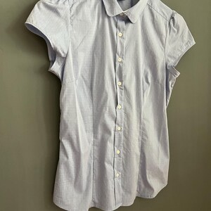 Op de afbeelding: Een lichtblauwe blouse met korte mouwen, een ronde kraag en een knoopsluiting. De blouse hangt aan een witte hanger tegen een grijze muur. De stof heeft een subtiel streepjespatroon.