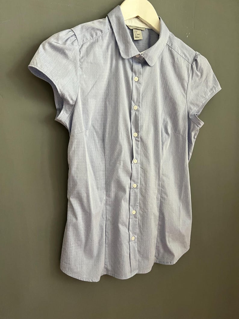 Op de afbeelding: Een lichtblauwe blouse met korte mouwen, een ronde kraag en een knoopsluiting. De blouse hangt aan een witte hanger tegen een grijze muur. De stof heeft een subtiel streepjespatroon.