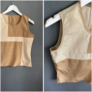 May include: A sleeveless top featuring a color-blocked design in shades of tan and cream. The garment is constructed with rectangular panels of fabric sewn together, creating a geometric pattern across the front, finished with a round neckline.