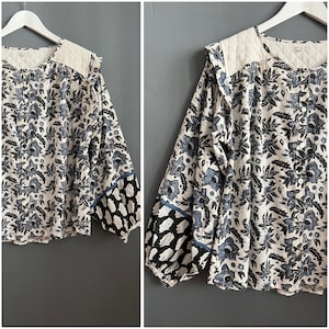 May include: A floral patterned blouse in blue and white, with long sleeves and a quilted yoke. The cuffs have a black and white design. The blouse is buttoned down the front and is displayed on a white hanger.