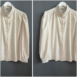 Puede incluir: Una blusa color crema con mangas largas y cuello alto, colgada de una percha blanca. La blusa tiene una botonadura frontal y detalles fruncidos en los hombros, creando un efecto de manga abullonada. La tela parece ligera.