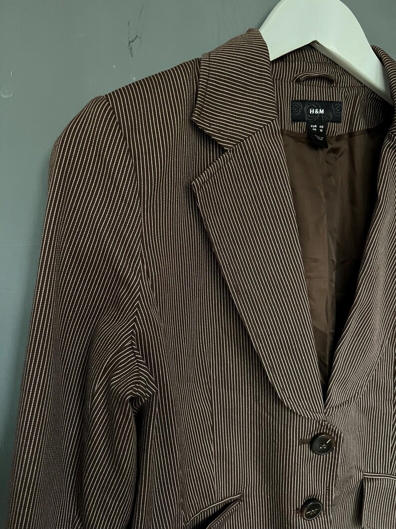 K&ouml;nnte beinhalten: Ein brauner Blazer mit wei&szlig;en Nadelstreifen und einem einzigen Knopfverschluss. Der Blazer hat einen Schalkragen und zwei Pattentaschen. Das Etikett auf dem Blazer lautet "H&M".