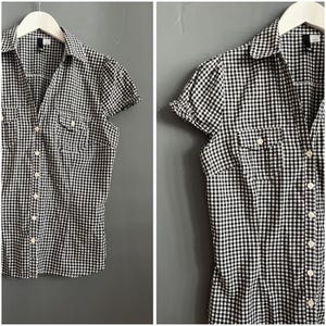 May include: A black and white gingham short-sleeved shirt with a button-down front and two chest pockets. The shirt is hanging on a white hanger against a grey background. The collar is pointed and the sleeves are slightly puffed.