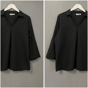 以下が含まれることがあります： Vネックカラーの黒い長袖シャツ。 シャツは白いハンガーにかけられており、背景はグレーです。 袖は七分丈です。