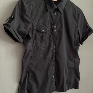 May include: Black short-sleeved button-down shirt with two chest pockets and roll-up sleeves. The shirt has a collared neckline and a small logo tag. The fabric appears to be slightly textured, with subtle embroidered details near the pockets.