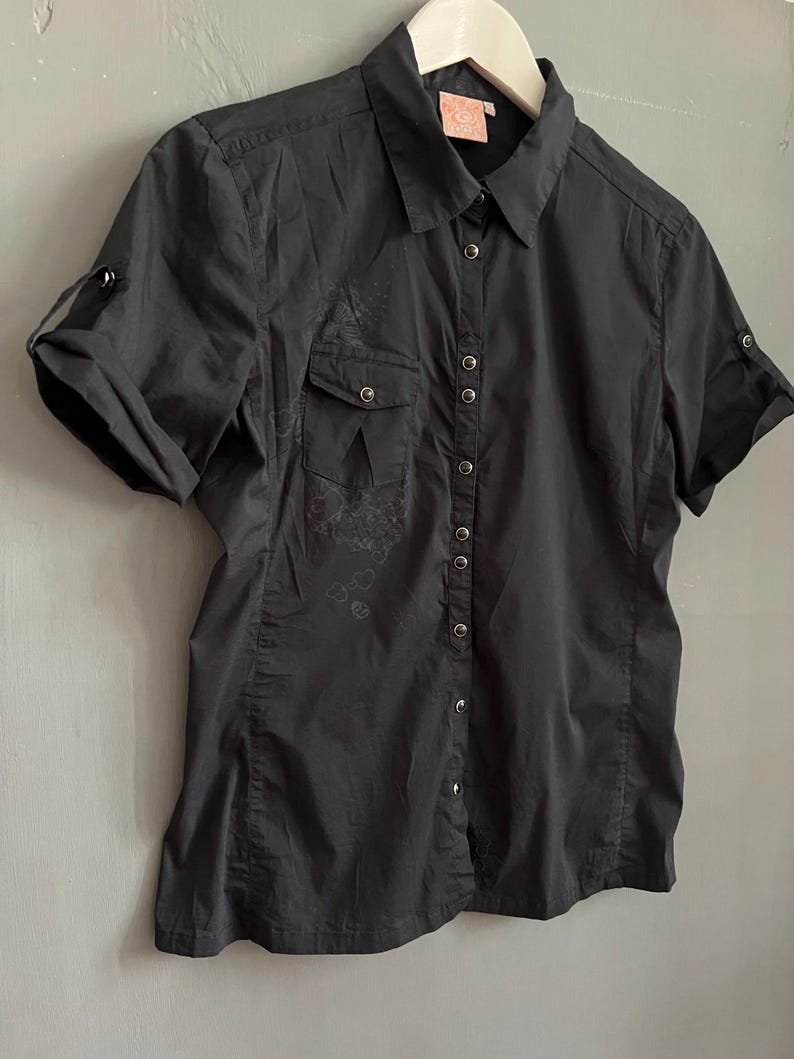 May include: Black short-sleeved button-down shirt with two chest pockets and roll-up sleeves. The shirt has a collared neckline and a small logo tag. The fabric appears to be slightly textured, with subtle embroidered details near the pockets.