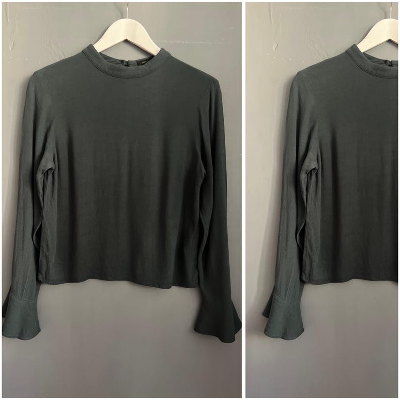 May include: A dark green long-sleeved blouse with a high neckline and flared sleeves. The blouse is hanging on a white hanger against a gray background. The sleeves have a bell-like shape at the wrist.