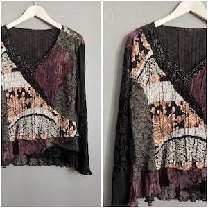 May include: A long-sleeved patchwork top with a V-neck. The top features a mix of textures and patterns, including floral prints, lace, and pleated fabrics in shades of black, burgundy, and cream. The top is hanging on a white hanger.