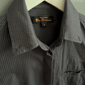 May include: A close-up of a charcoal grey button-down shirt with vertical stripes. The shirt features a collar, a chest pocket, and a tag reading "THE ORIGINAL Ben Sherman" with a size "S". The shirt is hanging on a white hanger.