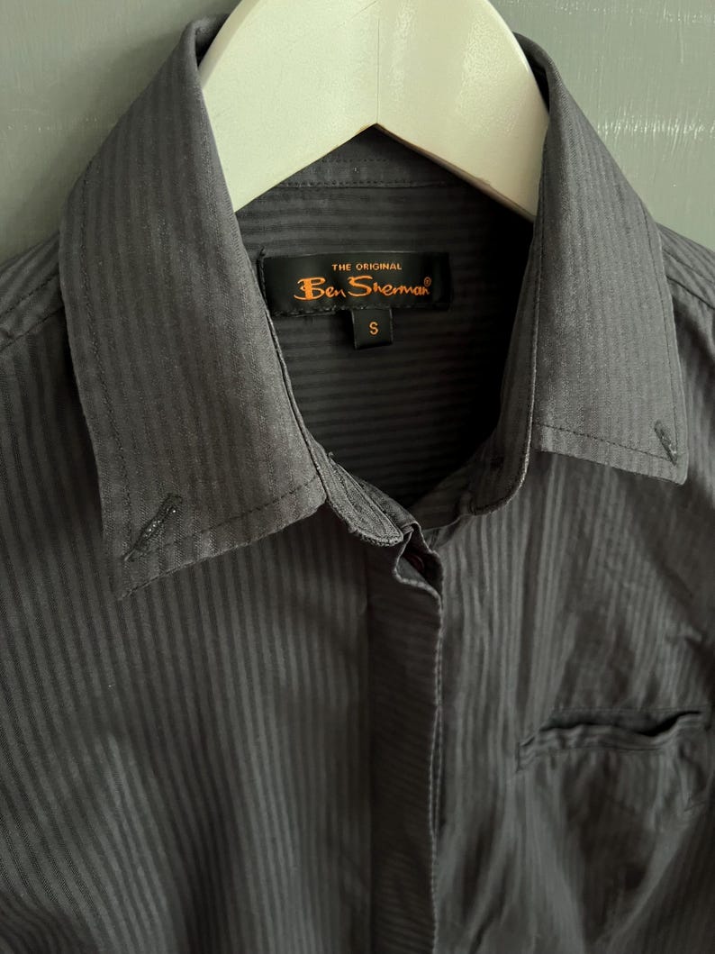 May include: A close-up of a charcoal grey button-down shirt with vertical stripes. The shirt features a collar, a chest pocket, and a tag reading "THE ORIGINAL Ben Sherman" with a size "S". The shirt is hanging on a white hanger.
