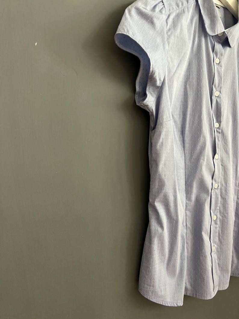 Op de afbeelding: Lichtblauw overhemd met knopen en korte, gefranjerde mouwen. Het overhemd heeft een subtiel stippenpatroon en een ronde kraag. De stof lijkt licht van gewicht, geschikt voor warmer weer. Het overhemd is aan de voorkant dichtgeknoopt.