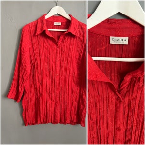 May include: A red, long-sleeved button-down shirt with a pleated design. The shirt has a collar and a label that reads "CANDA Collection of CA"