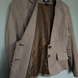 K&ouml;nnte beinhalten: Ein brauner Blazer mit wei&szlig;en Streifen, braunem Futter und zwei Kn&ouml;pfen. Der Blazer hat eine Tasche auf der linken Seite und das Etikett "H&M" ist innen sichtbar.