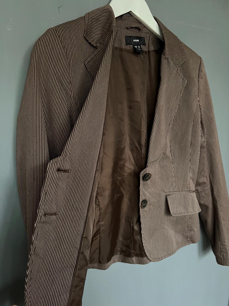 K&ouml;nnte beinhalten: Ein brauner Blazer mit wei&szlig;en Streifen, braunem Futter und zwei Kn&ouml;pfen. Der Blazer hat eine Tasche auf der linken Seite und das Etikett "H&M" ist innen sichtbar.