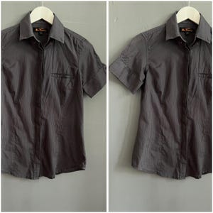 May include: A grey short-sleeved shirt with vertical stripes. The shirt features a chest pocket and is suspended on a white hanger. The collar is buttoned, and the sleeves are cuffed.