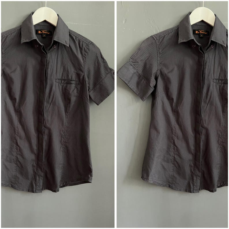 May include: A grey short-sleeved shirt with vertical stripes. The shirt features a chest pocket and is suspended on a white hanger. The collar is buttoned, and the sleeves are cuffed.