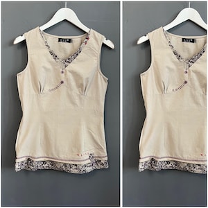 May include: A sleeveless beige top with a V-neck and decorative floral trim. The top features a button closure and a patterned hemline. The brand label "L33" is visible. The top is hanging on a white hanger against a grey background.