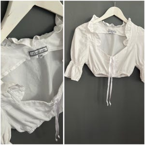 May include: White cropped blouse with ruffled neckline and short, puffed sleeves. The blouse has a square neckline and a drawstring closure. The label reads "dirndl.com" and size 34. The blouse is hanging on a white hanger against a gray background.