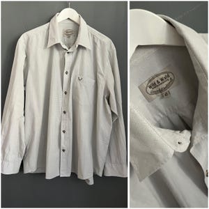 Puede incluir: Camisa abotonada gris claro con mangas largas, cuello y bolsillo en el pecho. La camisa tiene un pequeño diseño bordado en el bolsillo y una etiqueta que dice "Wild & Wald". La camisa está colgada en una percha blanca.