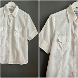 Può includere: Camicia bianca a maniche corte con due tasche sul petto. La camicia ha un colletto classico ed è realizzata in un tessuto leggero. La camicia è appesa a una gruccia bianca su sfondo grigio.