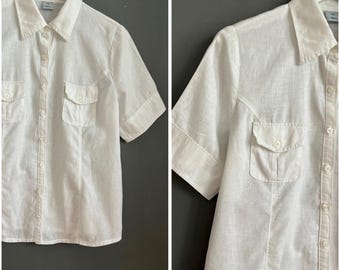 May include: White short-sleeve button-down shirt with two chest pockets. The shirt has a classic collar and is made of a lightweight fabric. The shirt is hanging on a white hanger against a gray background.