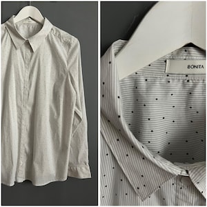 Puede incluir: Camisa blanca de botones con un pequeño patrón de lunares negros. La camisa tiene manga larga y cuello. La camisa tiene una etiqueta que dice "BONITA".