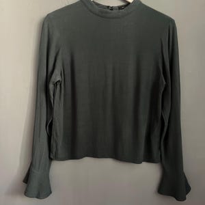 May include: A dark green long-sleeve blouse with a high neckline and flared sleeves. The blouse is hanging on a wooden hanger against a gray wall. The fabric appears to be a soft, flowing material.
