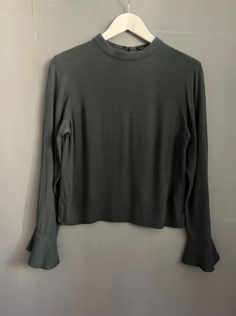 May include: A dark green long-sleeve blouse with a high neckline and flared sleeves. The blouse is hanging on a wooden hanger against a gray wall. The fabric appears to be a soft, flowing material.