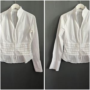 May include: A white long-sleeved shirt with a high collar and a full-length zipper. The shirt features a pleated design across the midsection and cuffs on the sleeves. The garment is hanging on a white hanger against a gray background.