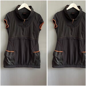 May include: A black short-sleeved tunic featuring a high collar with a front zipper, orange trim on the sleeves and pockets, and decorative stitching across the chest and hem. The garment includes two front pockets and a gathered bottom hem.