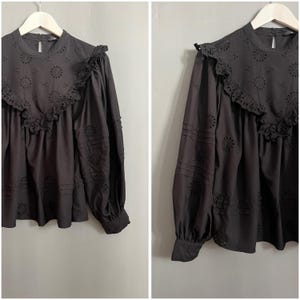 Puede incluir: Blusa negra de manga larga con cuello redondo y ribete de encaje. La blusa tiene un delicado patrón de ojal en toda la prenda.