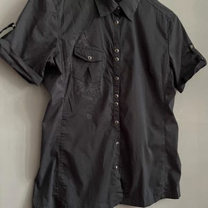 May include: A black short-sleeved button-down shirt with a collared neckline. The shirt features a chest pocket on the left side and decorative detailing. The sleeves can be rolled up and secured with a button. The shirt is hanging on a white hanger.